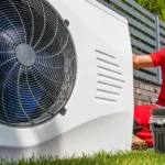 in Techniker in roter Arbeitskleidung und Kappe hockt im Freien neben einer weißen Wärmepumpe und greift in einen geöffneten Werkzeugkoffer.