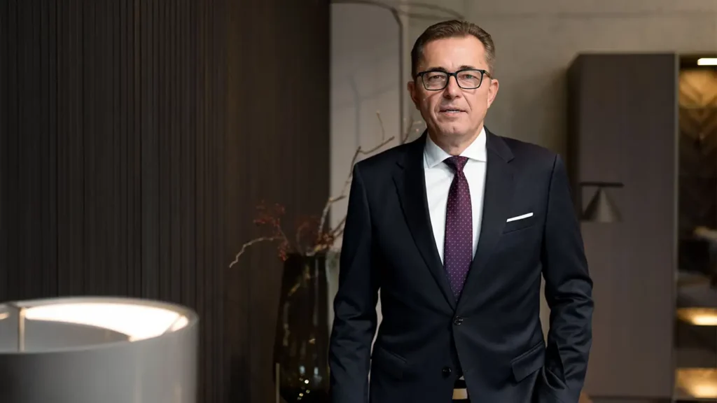 Porträt von ZVSHK-Präsident Michael Hilpert mit Brille, dunklem Anzug, weißem Hemd und gemusterter lila Krawatte in einem modern gestalteten Raum mit dunklen Wandpaneelen.