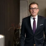 Porträt von ZVSHK-Präsident Michael Hilpert mit Brille, dunklem Anzug, weißem Hemd und gemusterter lila Krawatte in einem modern gestalteten Raum mit dunklen Wandpaneelen.